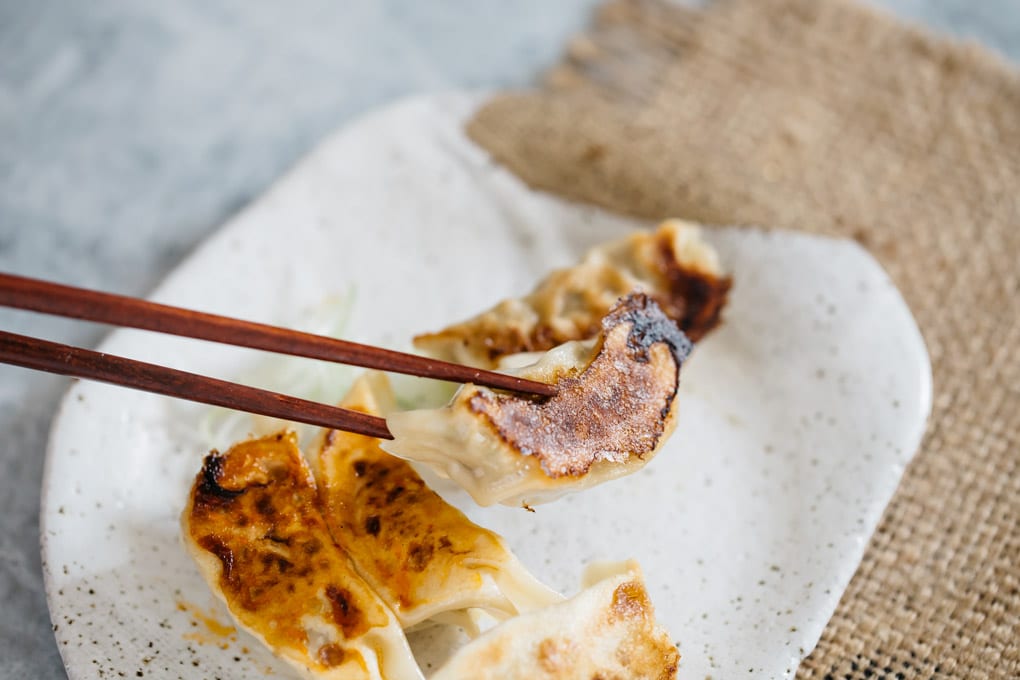 a pair of chopstick picking a piece of gyoza over a plate