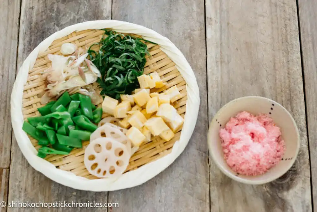 shiso, myoga, sugar snap peas, diced tamagoyaki, and rencon slices on a round bamboo tray and a small bowl of sakura denbu