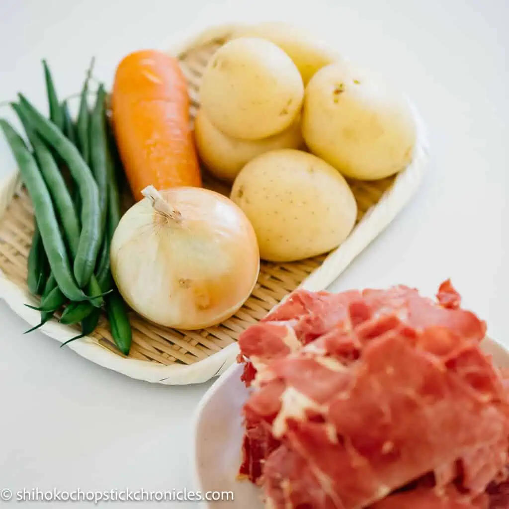 thinly sliced beef, potatoes, an onion, a carrot, and green beans on a bamboo tray