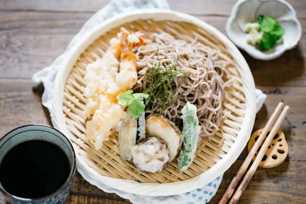 Tempura and soba noodles served together on a bamboo tray with garnish and dipping sauce in a small cup 