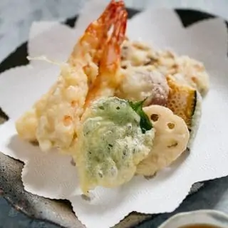 Two prawn tempura, shiso leaf tempura, renkon, kabocha and chikuwa fish cake tempura on a plate with a bowl of tentsuyu with gated daikon in it
