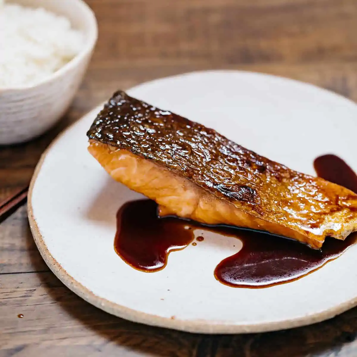 a piece of teriyaki glazed salmon on a round plate with a small bowl of rice in background
