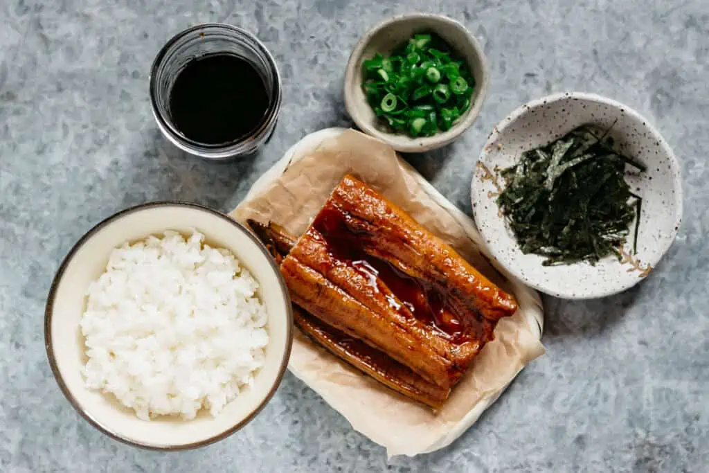 rice in a rice bowl, eel kabayaki on a bamboo tray, shredded nori sheet, unagi sauce, and chopped scallions