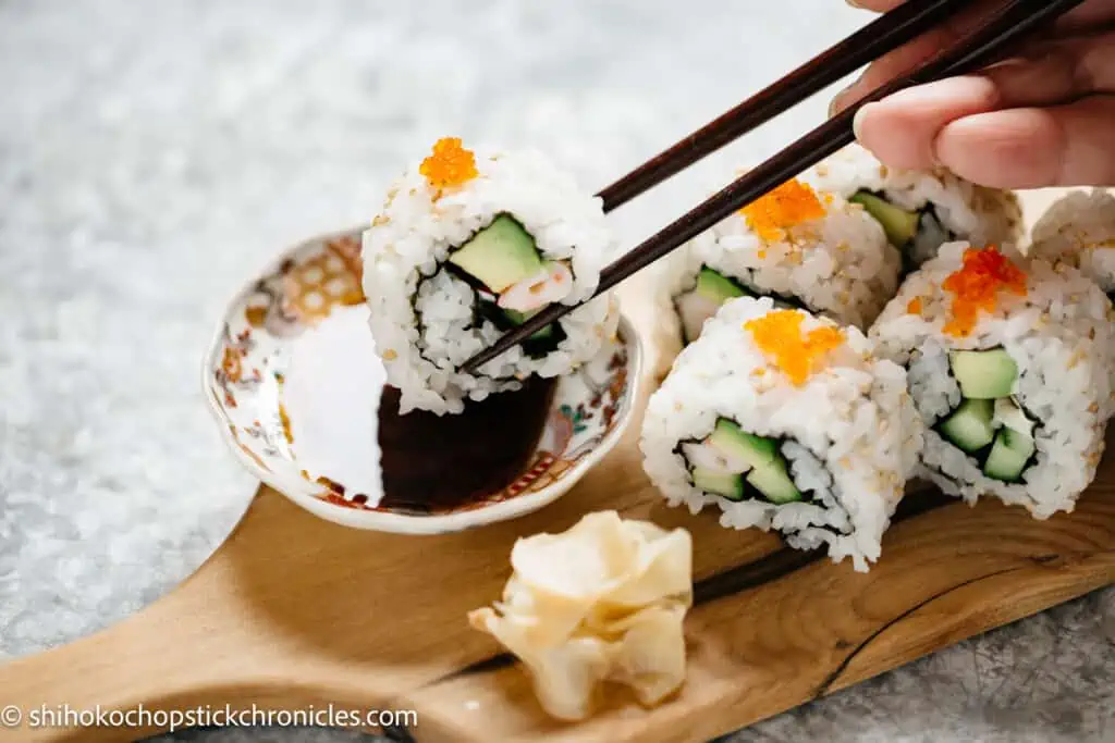 one piece of uramaki dipped in a small bowl of soy sauce by a pair of chopsticks